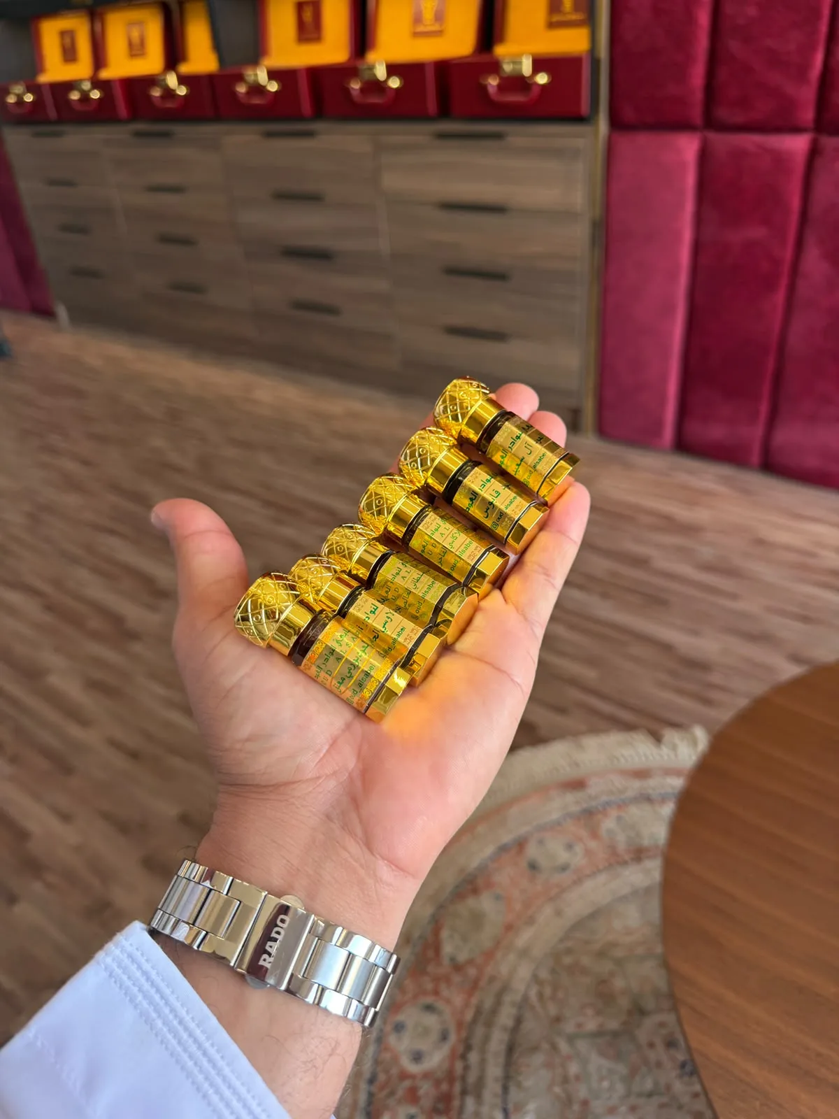 كفّ تحمل ست قوارير ذهبية صغيرة من دهن العود أمام أدراج المحل وصناديق الهدايا الحمراء